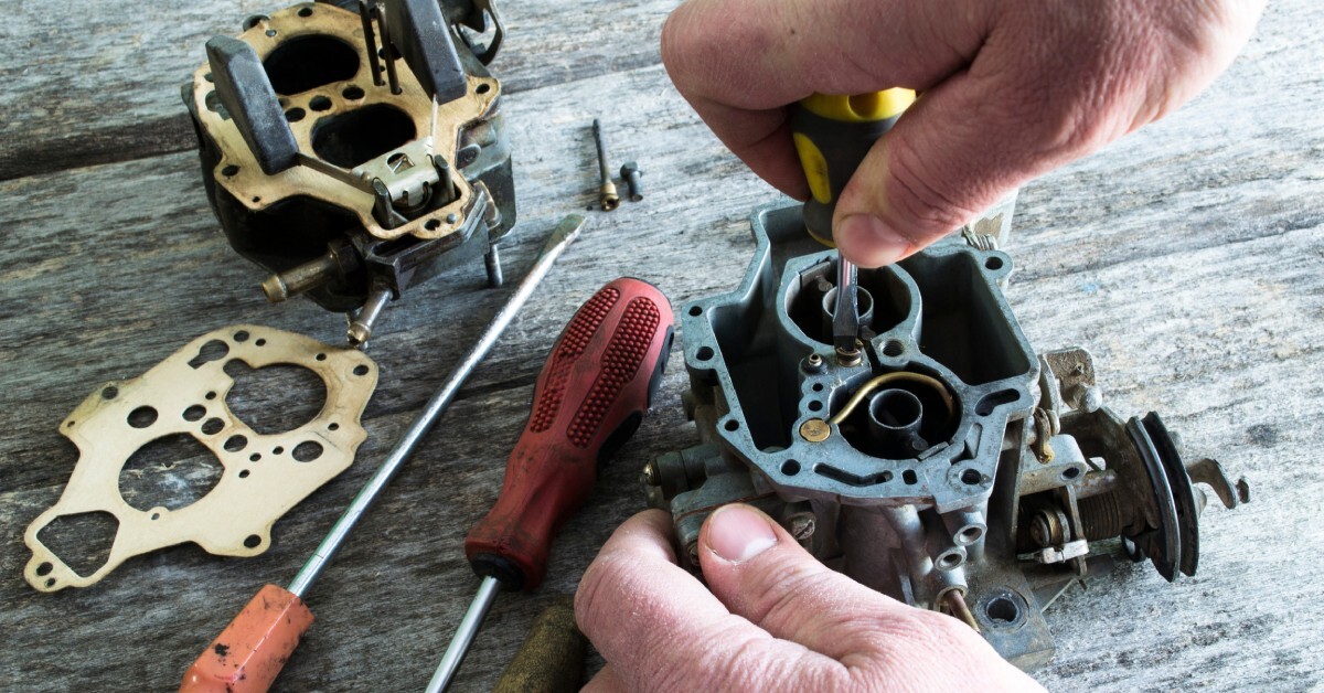 A close-up of a man's hands working on a carburetor with a screwdriver on a wooden table. Other screwdrivers lie nearby.