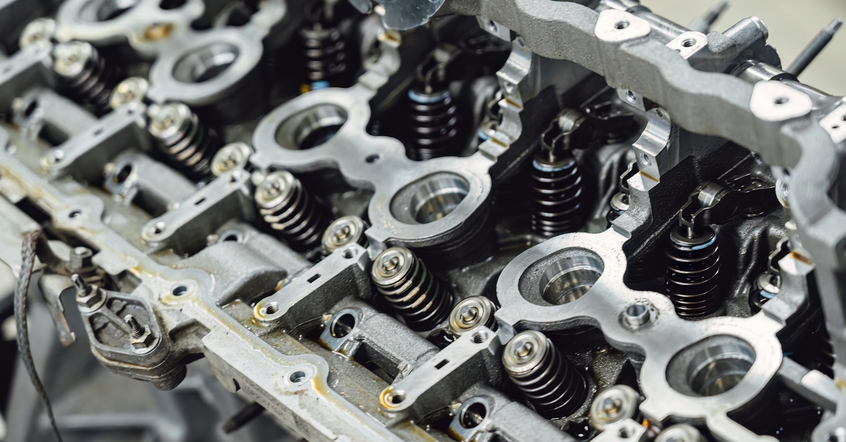 Close-up of a cylinder head of a car that is under repair. The block is silver and mostly clean, with a few drips of oil.