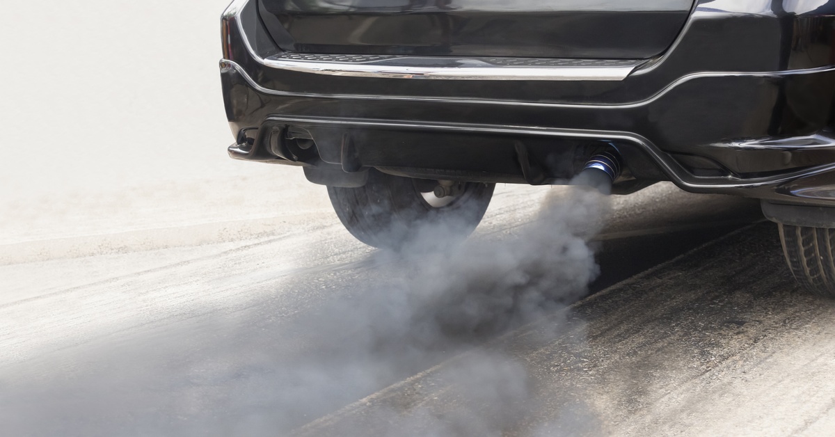 A car's tailpipe emits black and gray exhaust as it drives down a road. The vehicle is black and silver.