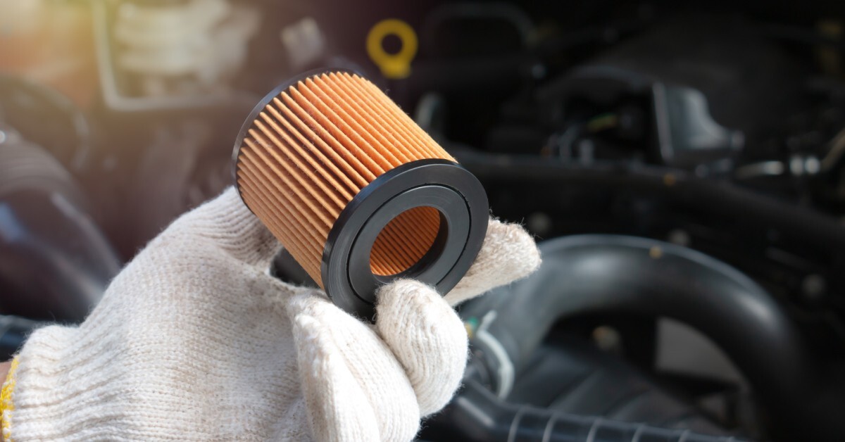 Close-up of a person's hand holding an orange and black fuel filter for an engine. The person wears a white glove..