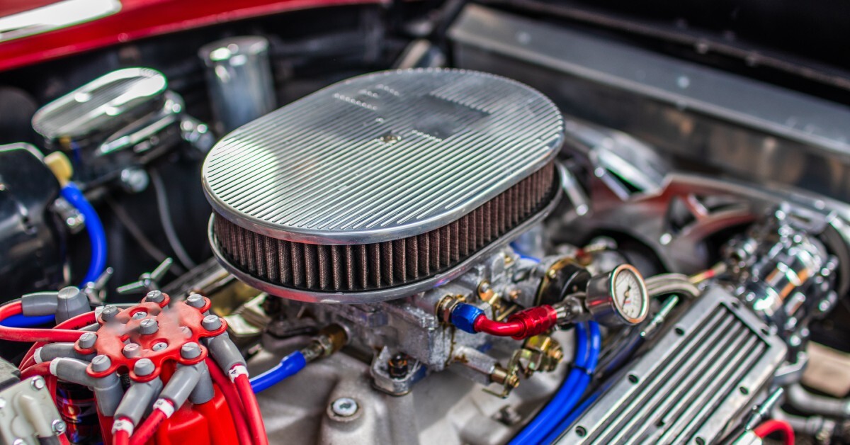 A shiny, silver air cleaner sits on top of a carburetor inside a vehicle's engine. The car has a red body.