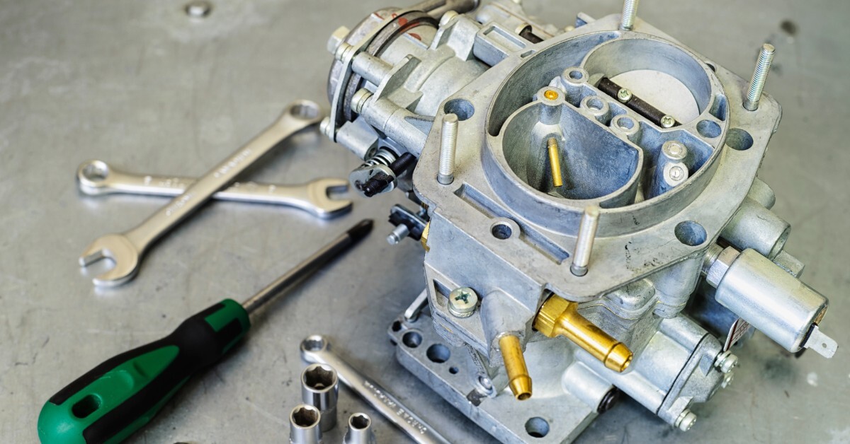 A new car carburetor sits on a silver counter. A green screwdriver and three silver wrenches lie next to it.