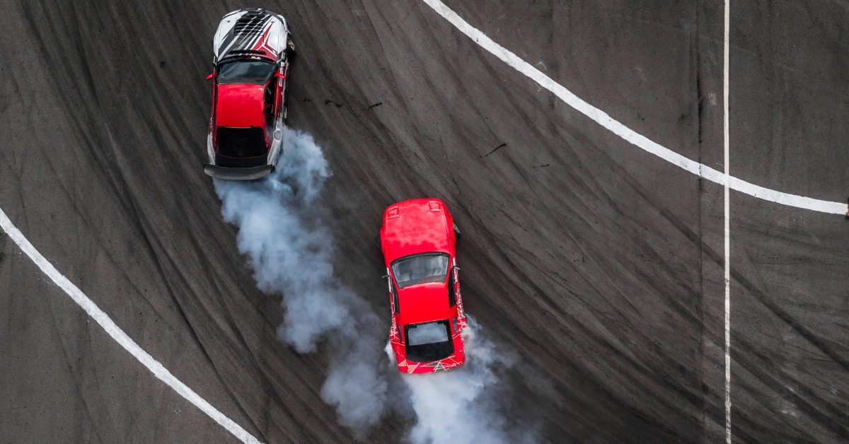 Overhead view of two red cars racing on a track with white lines. Smoke pours from the back of both vehicles.