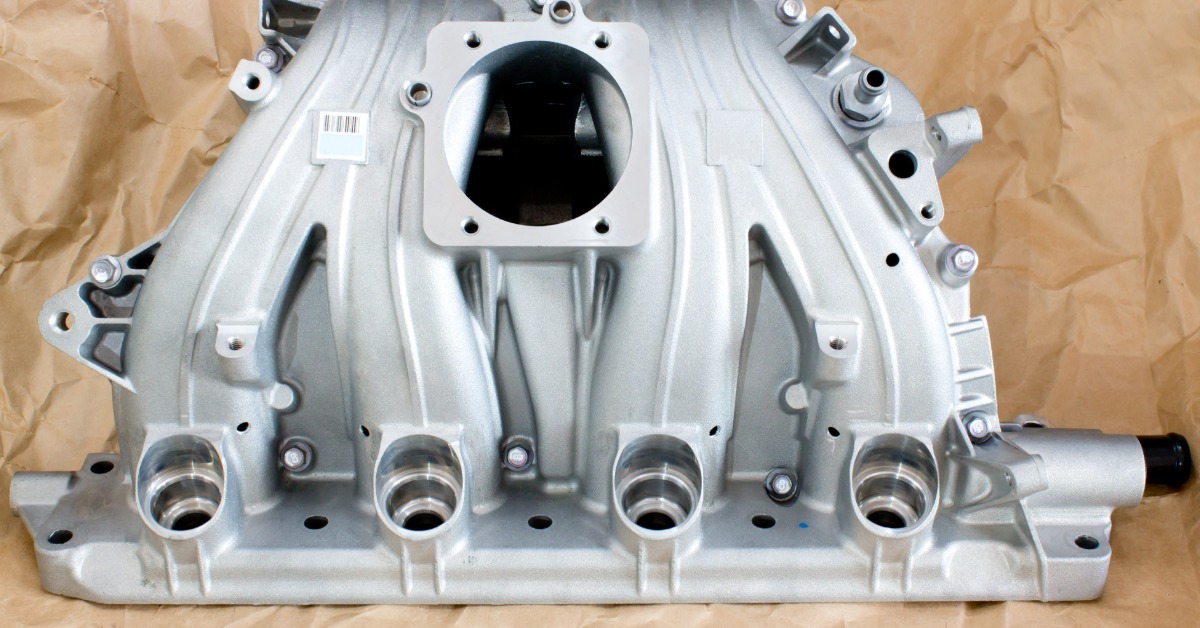 A brand-new, clean intake manifold sits on a surface covered with crinkly brown paper. The manifold features several holes.