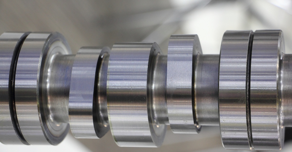 Close-up of the cams on a new camshaft for a vehicle. The shiny, silver component is against a gray background. Close-up of the cams on a new camshaft for a vehicle. The shiny, silver component is against a gray background.