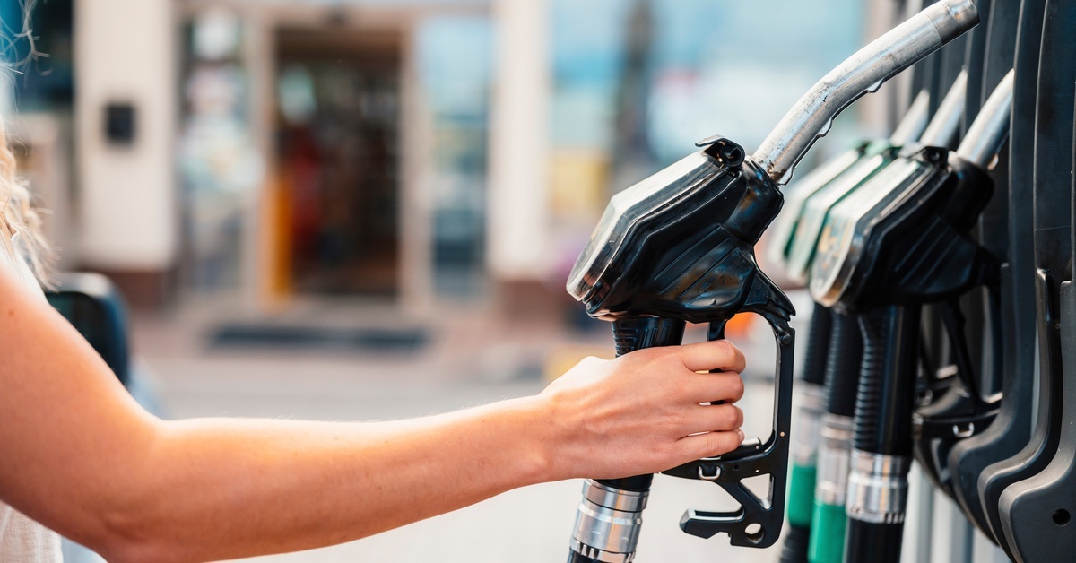 A person's hand reaches over and grabs a fuel pump at a gas station. The shop is visible in the background.